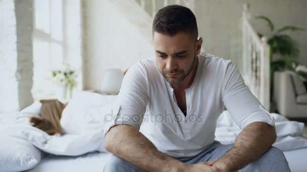 Расстроенный молодой человек, сидящий в постели, имеет проблемы с сексом, в то время как его девушка спит в спальне дома — стоковое видео