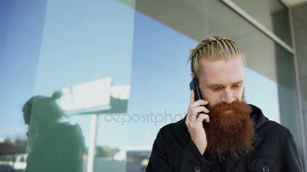 Fiatal szakállas csípő ember koncentrált beszél telefonon, a citystreet és beszélgetés közelében irodaház — Stock videók