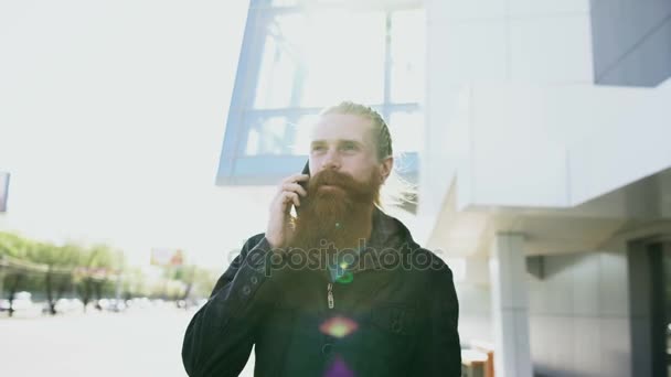 Dolly schot van jonge bebaarde hipster man geconcentreerd aan het praten over de telefoon op citystreet, gesprek in de buurt van kantoorgebouw — Stockvideo