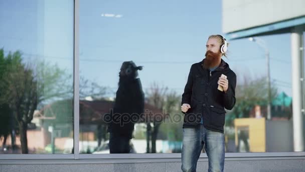 Gelukkig jonge bebaarde hipster man met hoofdtelefoon en smartphone luisteren naar muziek en dansen op straat stad — Stockvideo