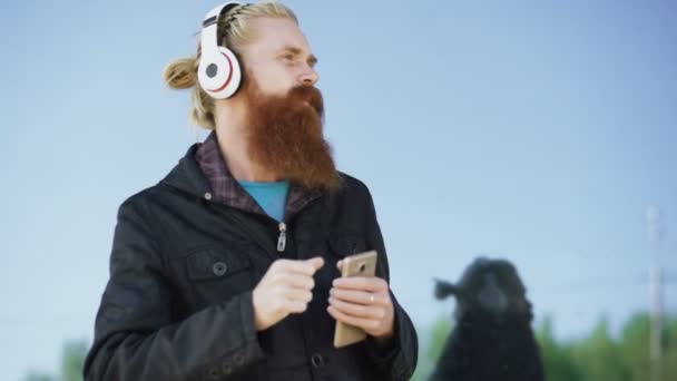 Happy young bearded hipster man with headphones and smartphone listen to music and dancing on city street — Stock Video