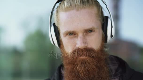 Retrato de primer plano de un joven hipster barbudo con auriculares escuchando música y sonriendo en la calle de la ciudad — Vídeos de Stock