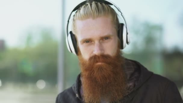 Retrato de primer plano de un joven hipster barbudo con auriculares escuchando música y sonriendo en la calle de la ciudad — Vídeo de stock