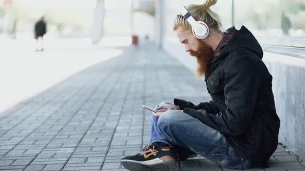 Young bearded hipster man with headphones sitting on road and using smartphone for listen to music and internet surfing outdoors at street — Stock Video