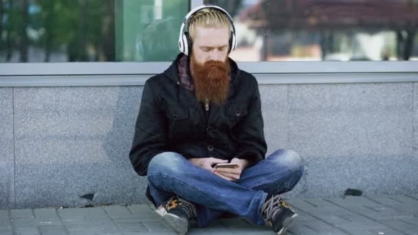 Ung skäggiga hipster mannen med hörlurar som sitter på vägen och använda smartphone för lyssna på musik och internetsurf utomhus på street — Stockvideo