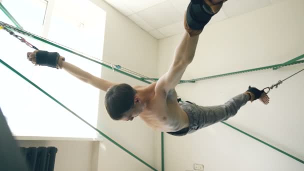 Молодой человек делает упражнения на четвертование оборудования для йоги и тянет свои руки и ноги веревками — стоковое видео