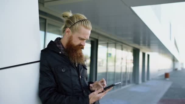 Joven barbudo hipster hombre usando teléfono inteligente fo Internet surf cerca de edificio de oficinas — Vídeo de stock