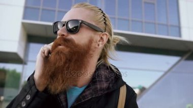 Genç gülümsemek gülümseyen ve ofis binaları yakınındaki smartphone konuşurken güneş gözlüğü erkekte hipster sakallı