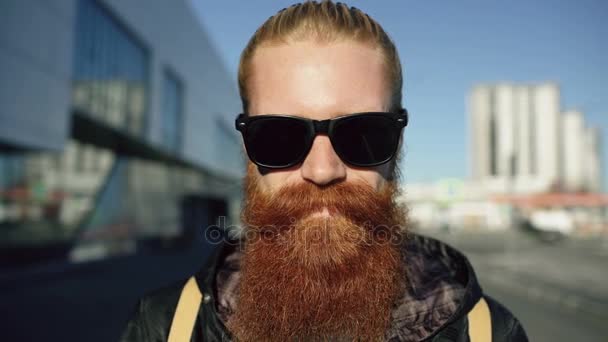 Portrait rapproché d'un jeune hipster barbu portant des lunettes de soleil souriant et posant alors qu'il se déplace dans la rue de la ville — Video