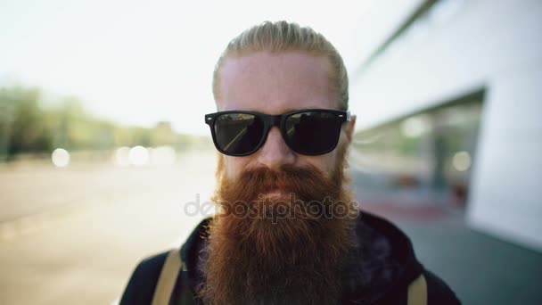Närbild porträtt av unga skäggiga hipster man i solglasögon leende och poserar resande stadsgata — Stockvideo