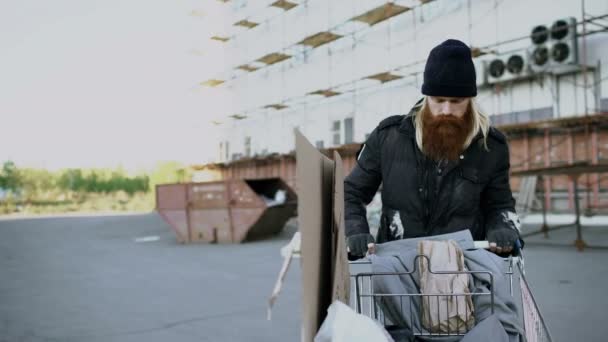 Dolly schot van bebaarde dakloze man lopend onderaan de straat met shooping kaart bij koude herfstdag — Stockvideo