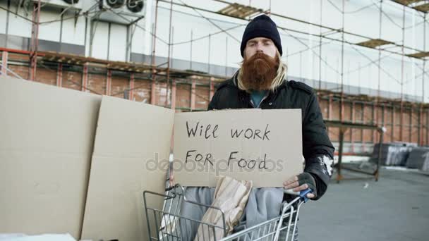 Ritratto di giovane senzatetto con cartone che guarda la macchina fotografica e vuole lavorare per il cibo in piedi vicino al carrello — Video Stock