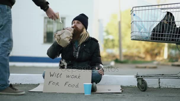 Young man helps to homeless person and giving him some food while beggar drink alcohol and sit near shopping cart at street — Stock Video