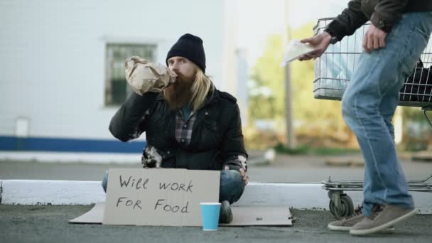 Giovane uomo aiuta a senzatetto e dandogli un po 'di cibo mentre mendicante bere alcol e sedersi vicino al carrello della spesa in strada — Video Stock
