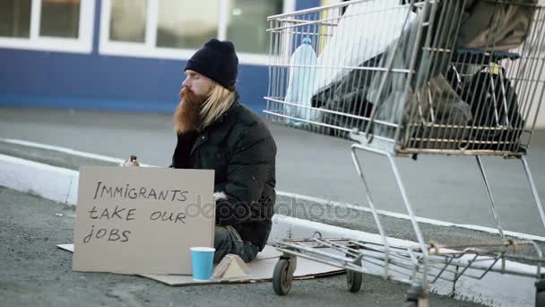 Hombre joven sin hogar barbudo con cartón sentado cerca del carrito de la compra y beber alcohol en el día frío. Crisis de los inmigrantes en Europa — Vídeos de Stock