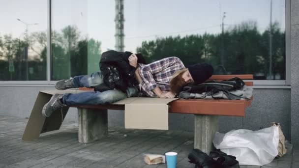 Hombre joven sin hogar tratando de dormir bajo la chaqueta en el banco en la acera — Vídeos de Stock