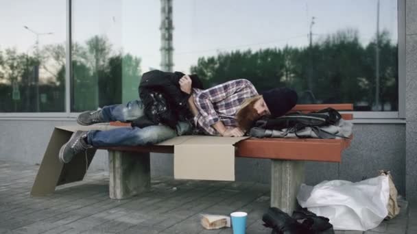 Young Homeless man trying to sleep under jacket on bench at the sidewalk — Stock Video