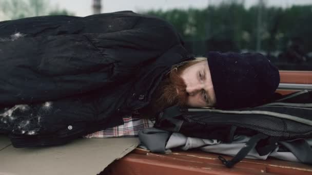 Молодой бездомный пытается спать под курткой на скамейке на тротуаре — стоковое видео