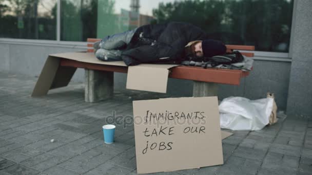 Hombre europeo sin hogar y sin trabajo con cartel de cartón duermen en el banco en la calle de la ciudad debido a la crisis de los inmigrantes en Europa — Vídeo de stock