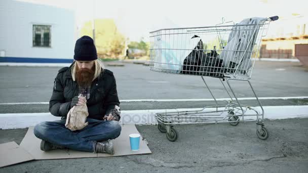 Бездомный молодой человек в грязной одежде пьет алкоголь, сидя возле тележки с покупками на улице в холодный зимний день — стоковое видео