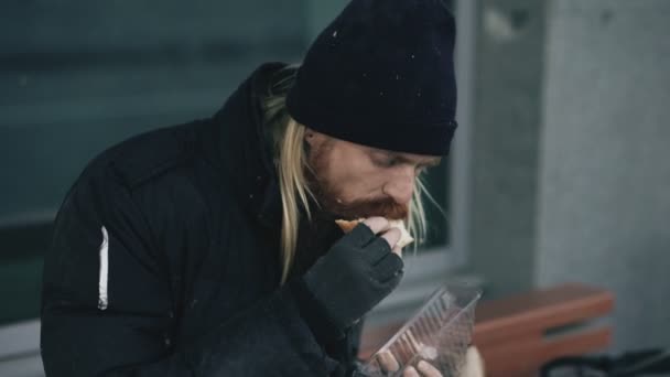 Hajléktalan fiatalember szendvicset eszik és iszik alkoholt papírzacskót padon: város utca, este — Stock videók