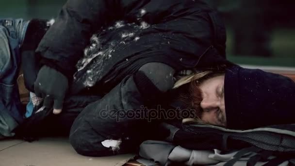 Молодой бездомный пьяница пытается спать на картонке на скамейке на тротуаре — стоковое видео