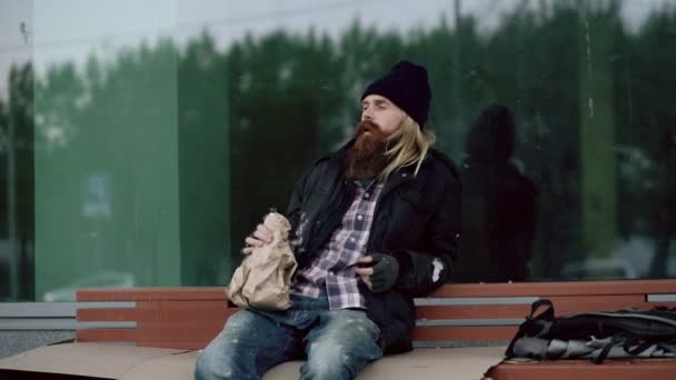 Пьяный бездомный молодой человек разговаривает с людьми, идущими рядом с ним и пьет алкоголь, сидя на скамейке на тротуаре — стоковое видео