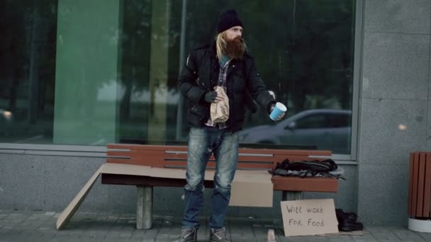 Дуже п'яний бездомний чоловік з картонним та алкогольним знаком благає грошей, стоячи біля лави на тротуарі — стокове відео