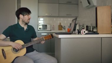 Neşeli genç adam evde dizüstü bilgisayar kullanan gitar çalmayı öğrenme mutfak oturma