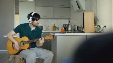 Evde dizüstü bilgisayar kullanan gitar çalmayı öğrenme mutfağa oturan kulaklıklar ile neşeli genç adam