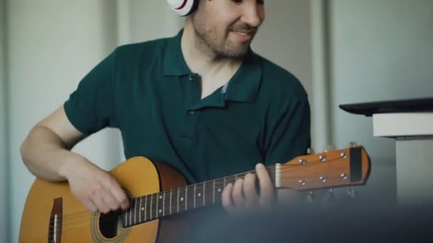 Cheerful young man with headphones sitting at kitchen learning to play guitar using laptop computer at home — Stock Video