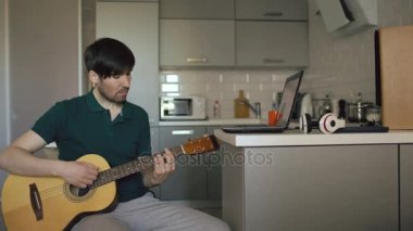 Çekici bir genç adam evde dizüstü bilgisayar kullanan gitar çalmayı öğrenme mutfak oturma
