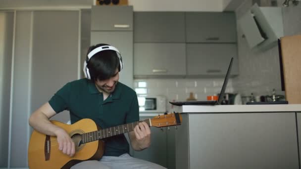 Joyeux jeune homme avec écouteurs assis à la cuisine apprenant à jouer de la guitare à l'aide d'un ordinateur portable à la maison — Video