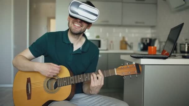 Feliz joven sentado en la cocina aprendiendo a tocar la guitarra con auriculares VR 360 y se siente guitarrista en concierto en casa — Vídeo de stock