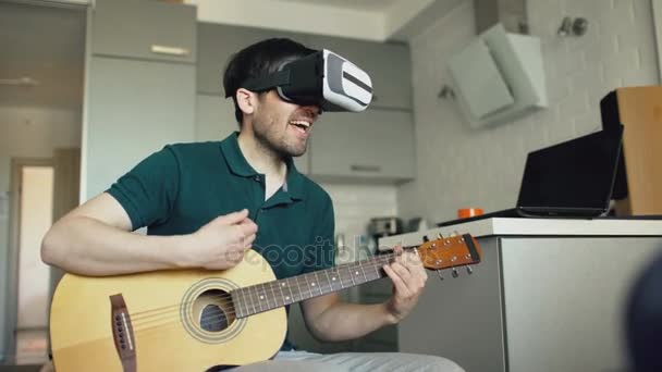 Jeune homme heureux assis à la cuisine apprenant à jouer de la guitare en utilisant VR 360 casque et se sent guitariste au concert à la maison — Video