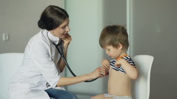 Mujer joven médico escuchando niño pequeño con estetoscopio en el consultorio médico — Vídeo de stock