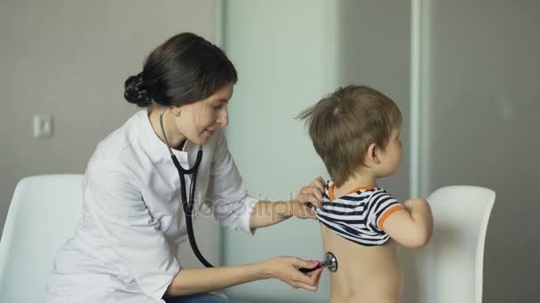Молодая женщина врач говорить маленький мальчик и слушать со стетоскопом в медицинском кабинете — стоковое видео