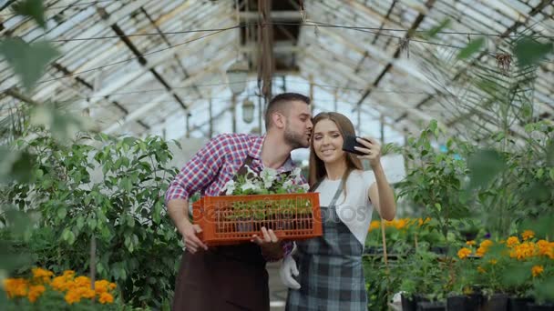 Alegre pareja cariñosa jardineros tomando foto selfie en la cámara del teléfono inteligente y besándose mientras trabaja en invernadero — Vídeos de Stock