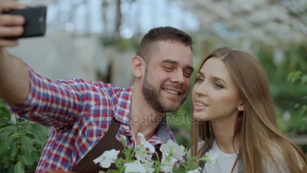 Веселая любящая пара садовников фотографирует селфи на камеру смартфона и целуется во время работы в оранжерее — стоковое видео