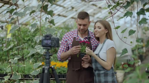 Joven blogger sonriente pareja jardineros en delantal sosteniendo flor hablando y grabando video blog para vlog en línea sobre jardinería — Vídeos de Stock