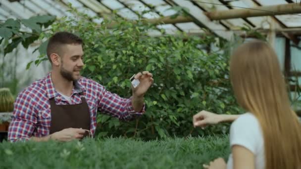 Jonge lachende verkoper bloemist werken in tuincentrum. Man geeft boodschappentas aan klant- en betaling maken per creditcard bij winkel — Stockvideo