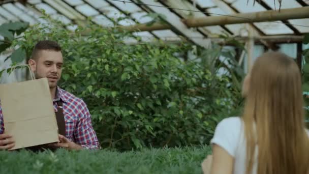 Joven florista vendedor sonriente trabajando en el centro del jardín. Hombre da bolsa de la compra al cliente y hacer el pago con tarjeta de crédito en la tienda — Vídeos de Stock