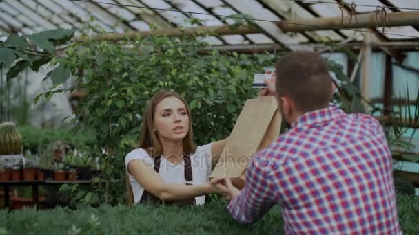 Joven florista vendedor sonriente trabajando en el centro del jardín. Mujer da bolsa de la compra al cliente y hacer el pago con tarjeta de crédito en la tienda — Vídeo de stock