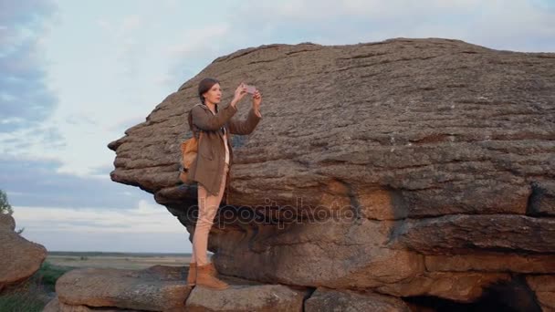 Junge Touristin fotografiert Landschaft mit ihrer Smartphone-Kamera nach einer Wanderung auf einem Felsen bei Sonnenuntergang — Stockvideo