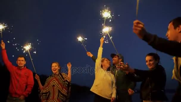 Группа молодых друзей устраивает пляжную вечеринку. Друзья танцуют и празднуют с бенгальскими огнями на закате — стоковое видео