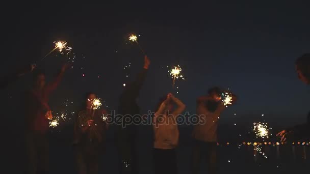 Group of young friends having a beach party. Friends dancing and celebrating with sparklers in twilight sunset — Stock Video
