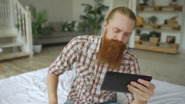 Jovem barbudo usando tablet computador ter vídeo chat sentado na cama em casa — Vídeo de Stock