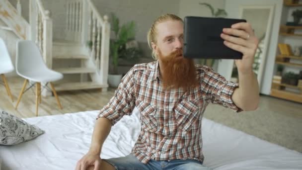 Młody Brodaty mężczyzna przy użyciu komputera typu tablet o wideo czat, siedząc na łóżku w domu — Wideo stockowe