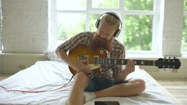 Homem barbudo atraente sentado na cama aprendendo a tocar guitarra usando o computador tablet no quarto moderno em casa — Vídeo de Stock