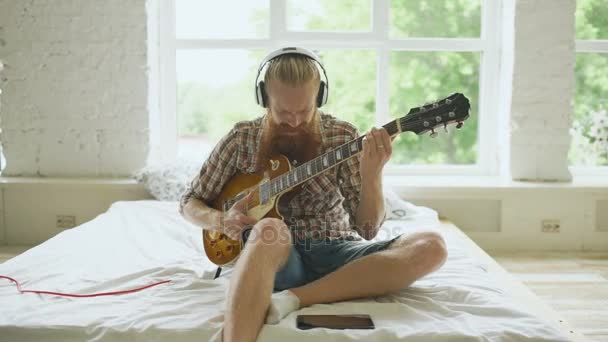 Attraente uomo barbuto in cuffia seduto sul letto imparare a suonare la chitarra utilizzando tablet computer in camera da letto moderna — Video Stock